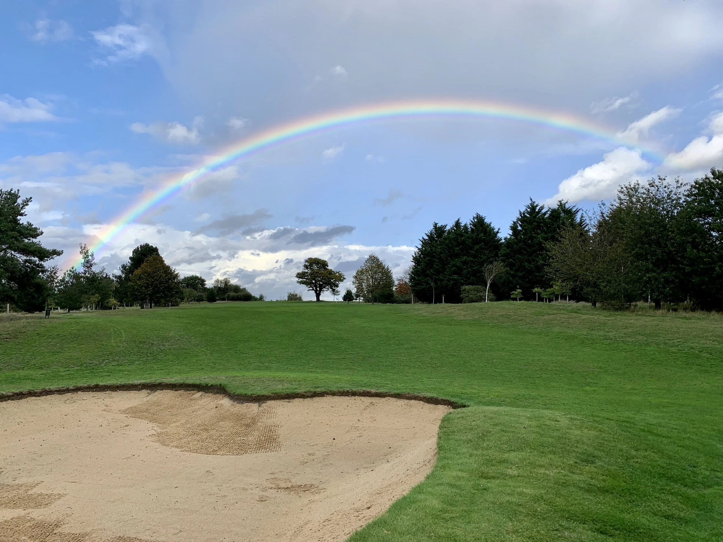 A golf course with a sand bunker in the foreground, green grass, trees in the background, and a rainbow in the sky.