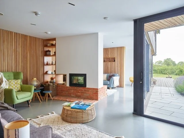 Modern living room with green armchair, gray sofa, woven coffee table, fireplace, and sliding glass door opening to outdoor patio with green landscape.