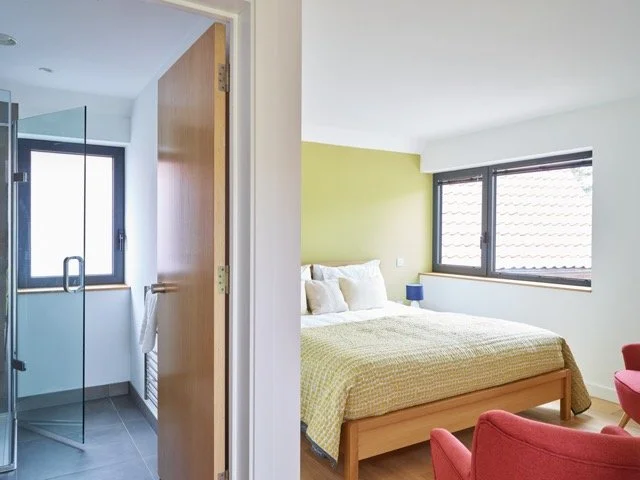 Split view of a bathroom with glass shower and a bedroom with bed, two windows, and red chairs.