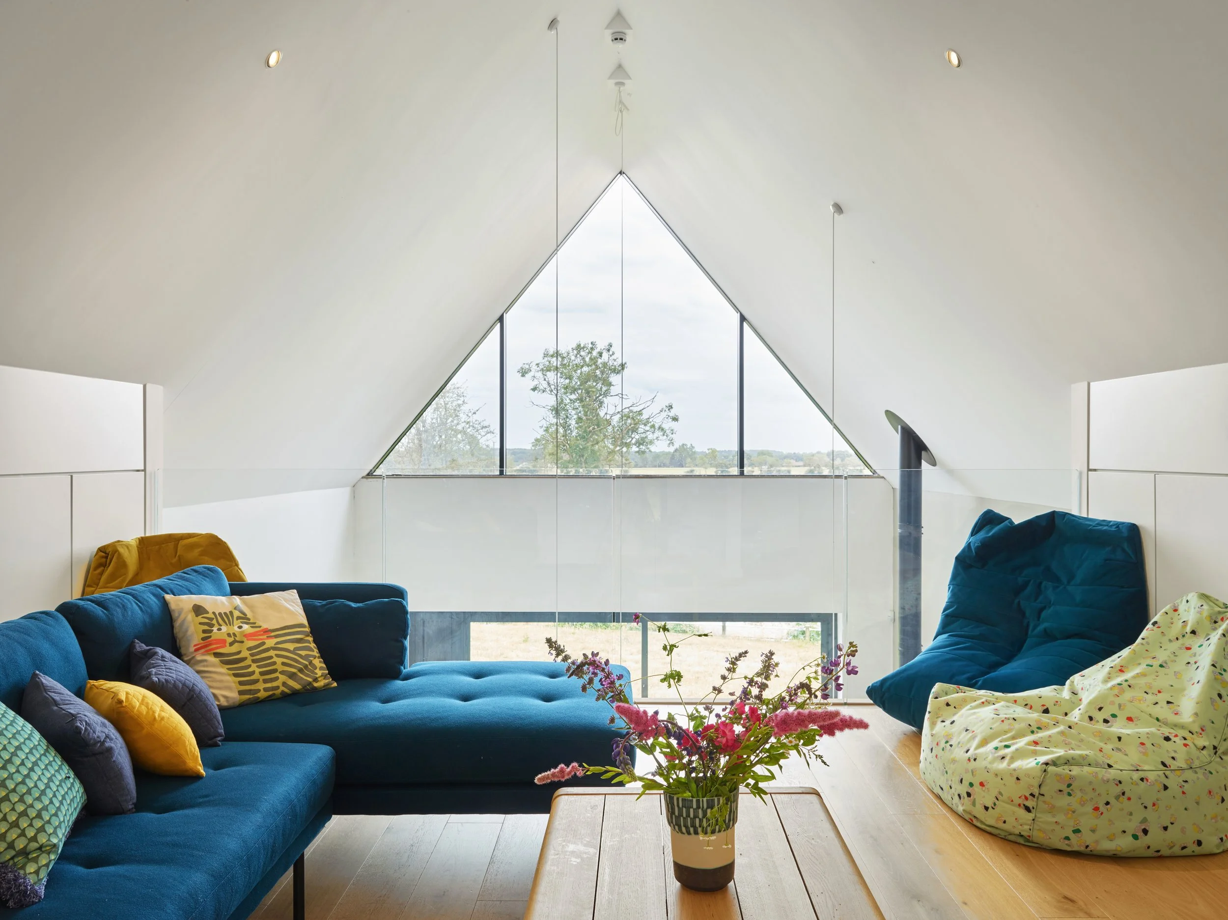 Interior of an attic living room with a sloped ceiling, large triangular window, blue sofa, colorful pillows, a yellow armchair, bean bag chairs, a wooden coffee table with a vase of pink and purple flowers, and a view of a tree outside.