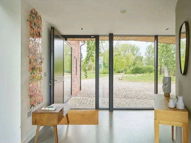 Interior of a modern home with large glass front door and windows, wooden furniture including a console table and side table, decorative vases, a colorful wall art piece, and a view of greenery outside.