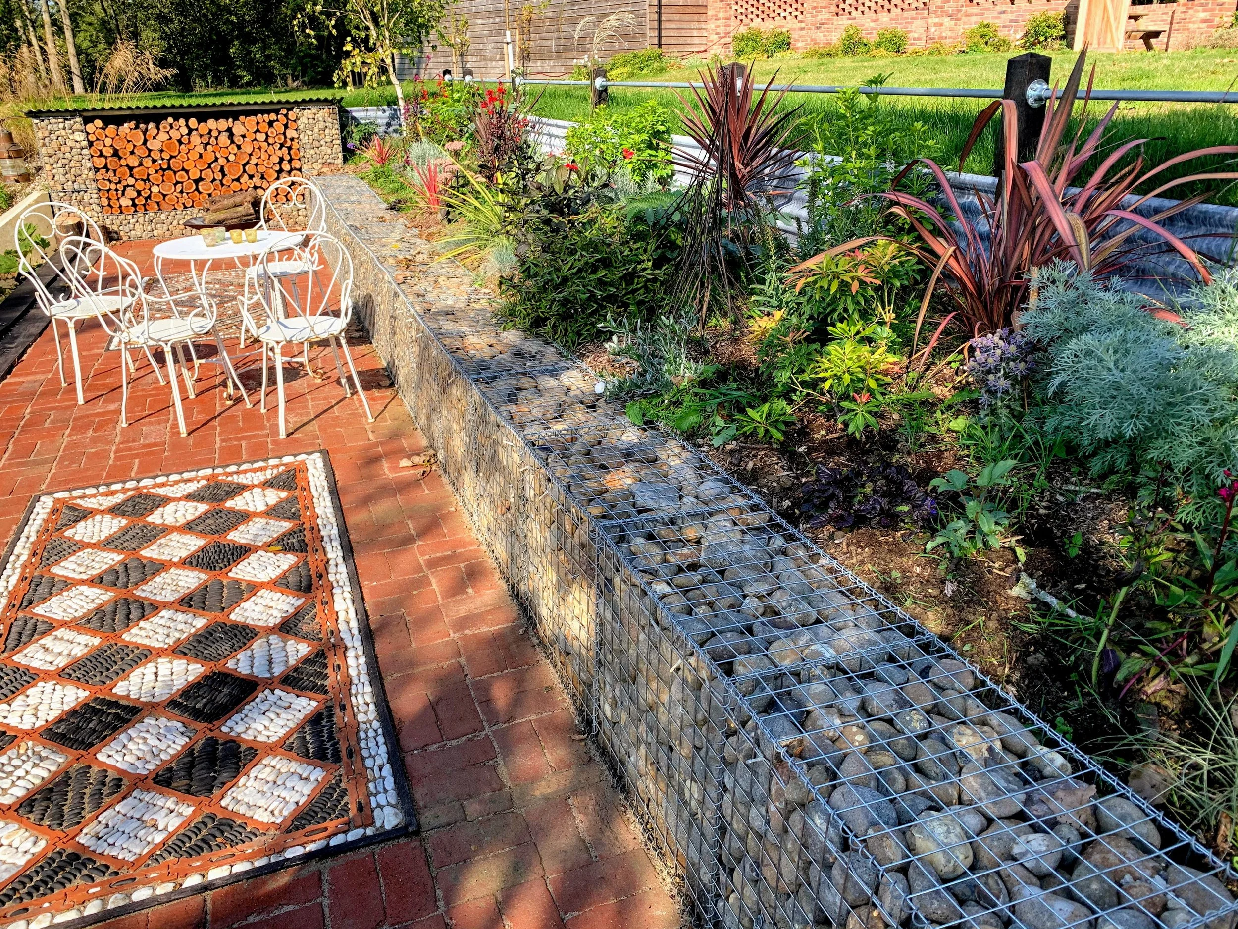 A garden patio with white metal chairs around a small table, a brick floor with an outdoor rug, a raised garden bed with various plants and a gabion wall with rocks and a wooden log storage area in the background.