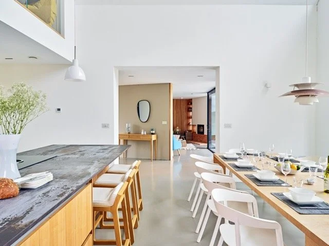 Modern dining area with a long wooden table set for a meal, white chairs, and a view into a living room with a TV and cozy seating.