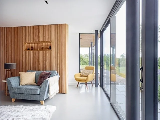 Modern living room with wooden accent wall, blue sofa with pillows, yellow armchair, floor lamp, and large glass sliding doors leading to an outdoor patio with greenery.