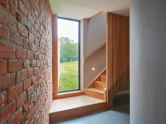 Modern stairway with wooden steps and railing inside a house, next to a large window looking out to greenery, with a brick wall and a gray wall interior.