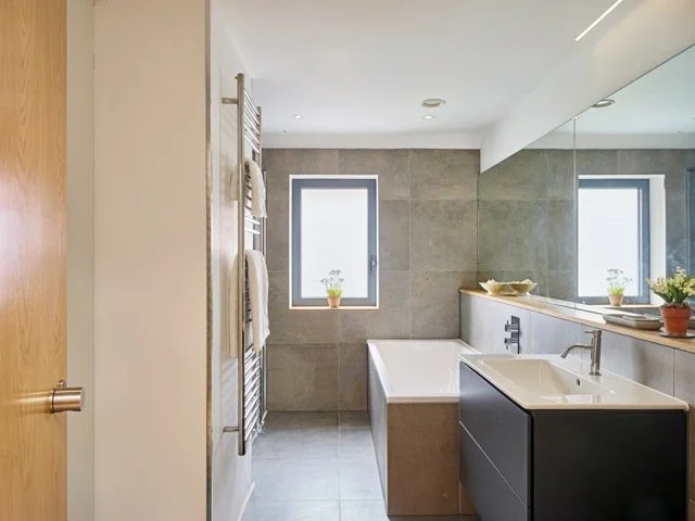 Modern bathroom with bathtub, sink, large mirror, and two small windows with potted plants.