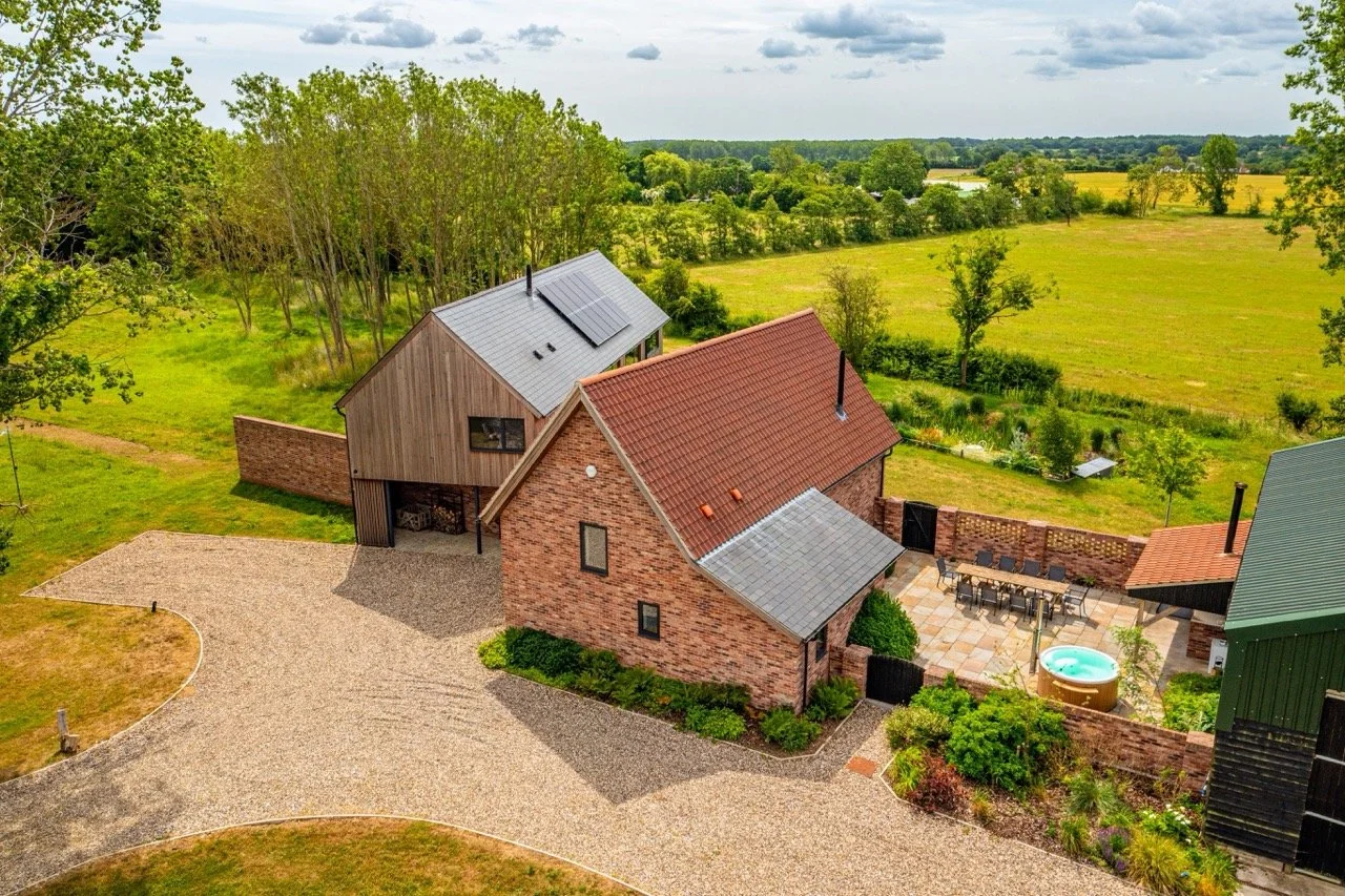 A rural house with a red brick exterior, gray and green metal roofs, a gravel driveway, and a backyard with a hot tub and patio furniture; surrounding greenery and open fields under a cloudy sky.