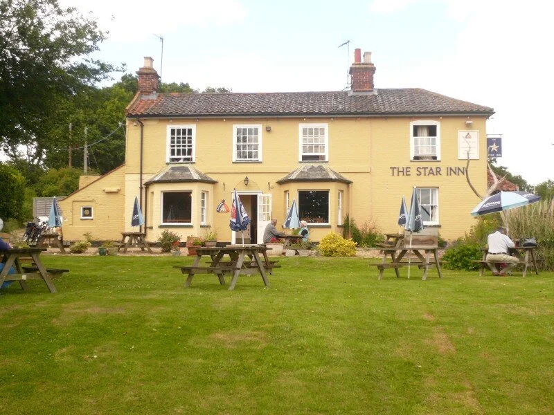Yellow two-story building with a sign that reads 'The Star Inn'. Outdoor seating with picnic tables and umbrellas, some people sitting and talking in the grassy area in front of the building.