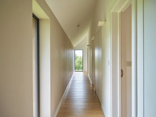 A bright, narrow hallway with light-colored walls, wooden flooring, and a door at the end with a window showing greenery outside.