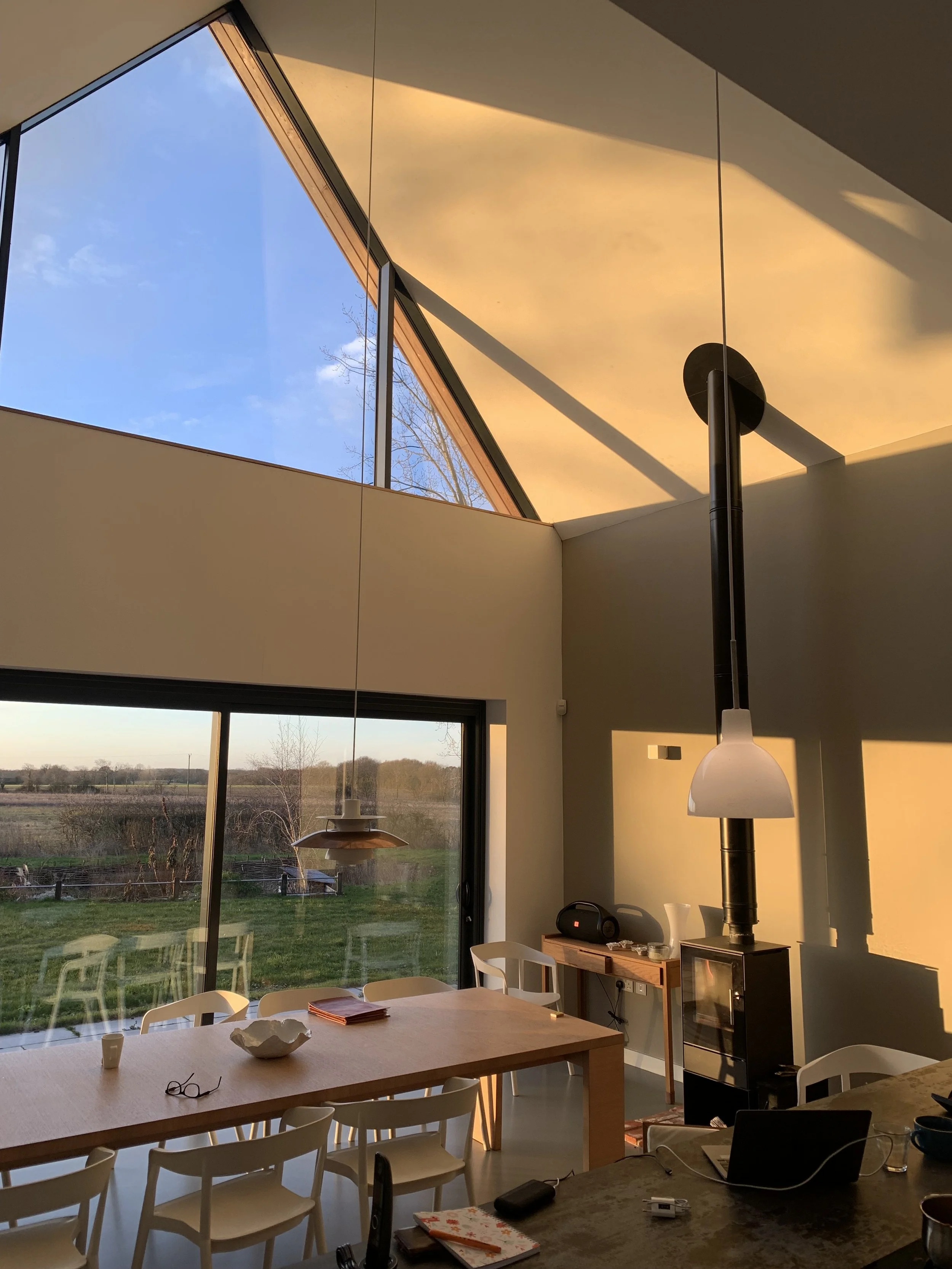 Interior of a modern dining room with large windows and a skylight showing a blue sky and trees outside. There is a wooden dining table with white chairs, a hanging lamp, and a small black stove or heater.