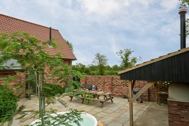 A backyard patio area with a wooden table and chairs, a Pizza oven with a tiled roof, a brick wall, and an outdoor wood-fired oven under a black roofed structure.