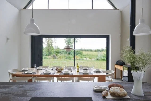 Modern dining room with large window overlooking green countryside, set with plates, glasses, and silverware, with white hanging pendant lights and a wooden sideboard.