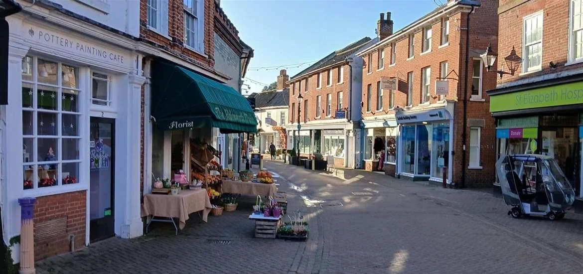 A small town street with shops, including a pottery painting cafe, a florist, and a pharmacy, with stalls of flowers outside.