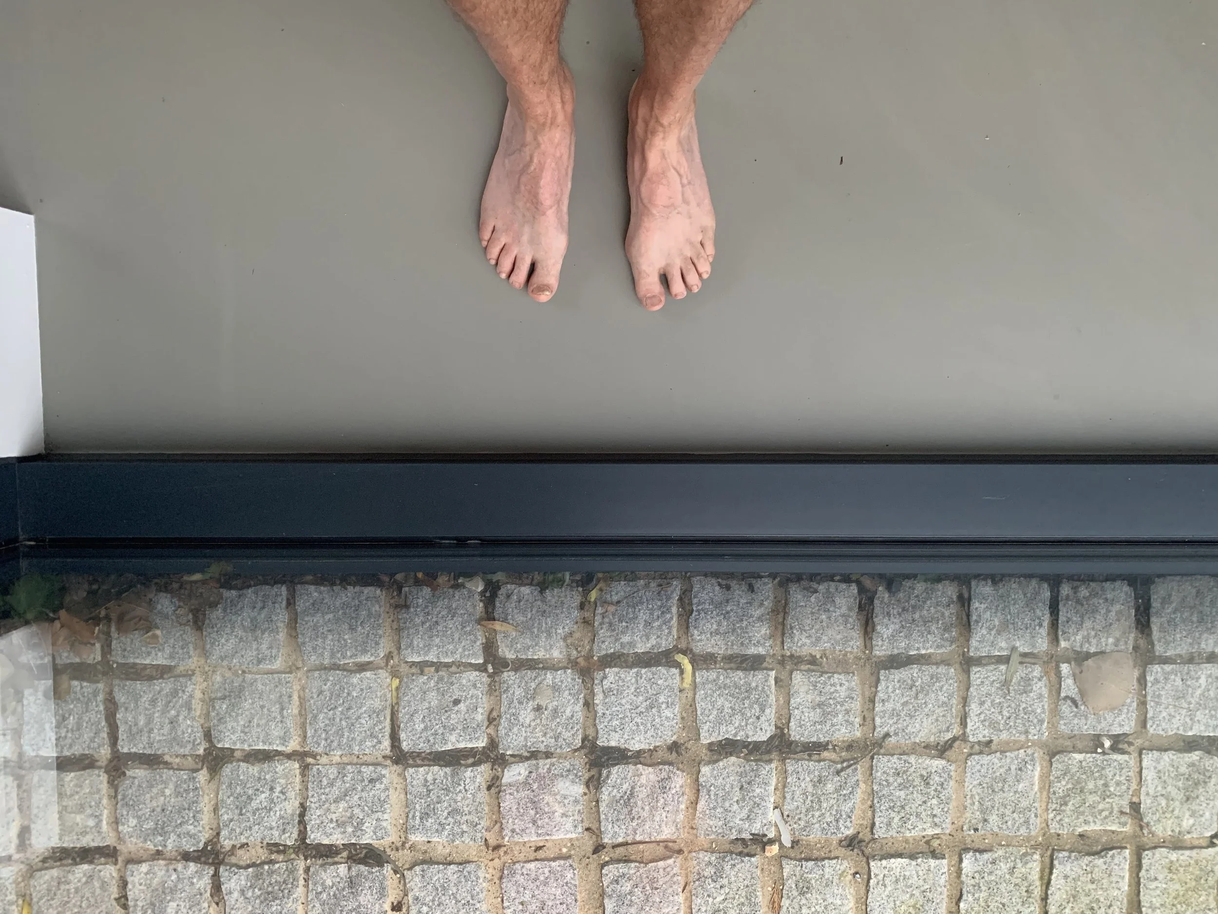 A top-down view of a person's bare feet standing on a gray floor next to a glass window with a cobblestone sidewalk outside.