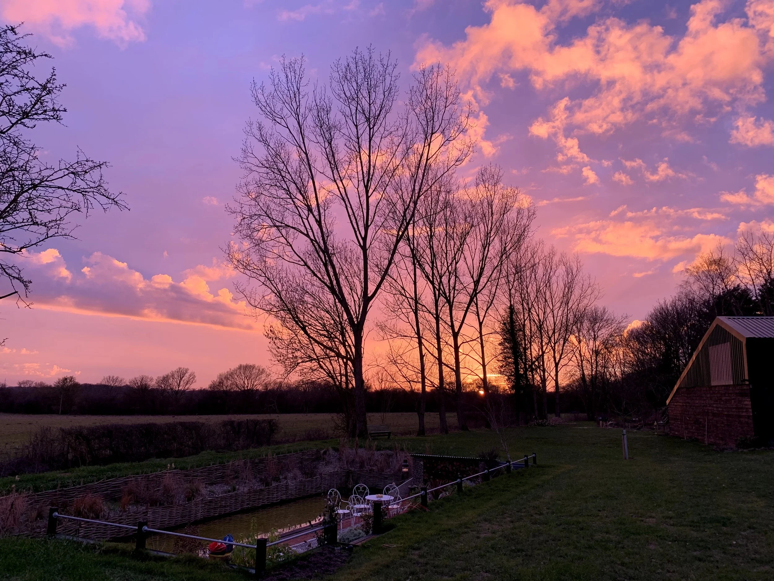 A rural landscape at sunset with colorful sky, bare trees, a small pond, and a brick barn on the right.
