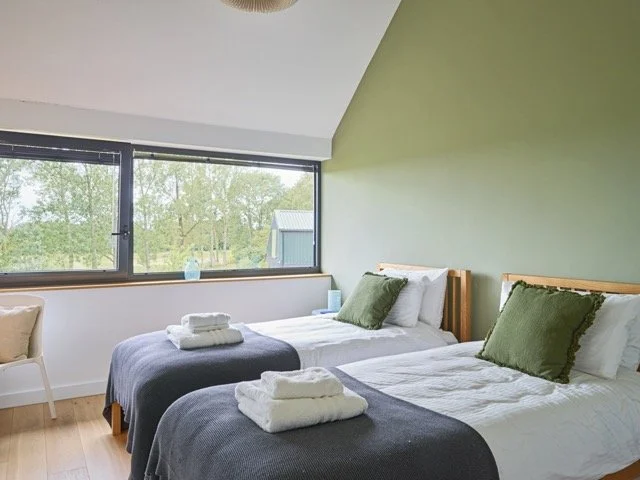 A bedroom with two single beds, green accent wall, and large windows showing trees outside