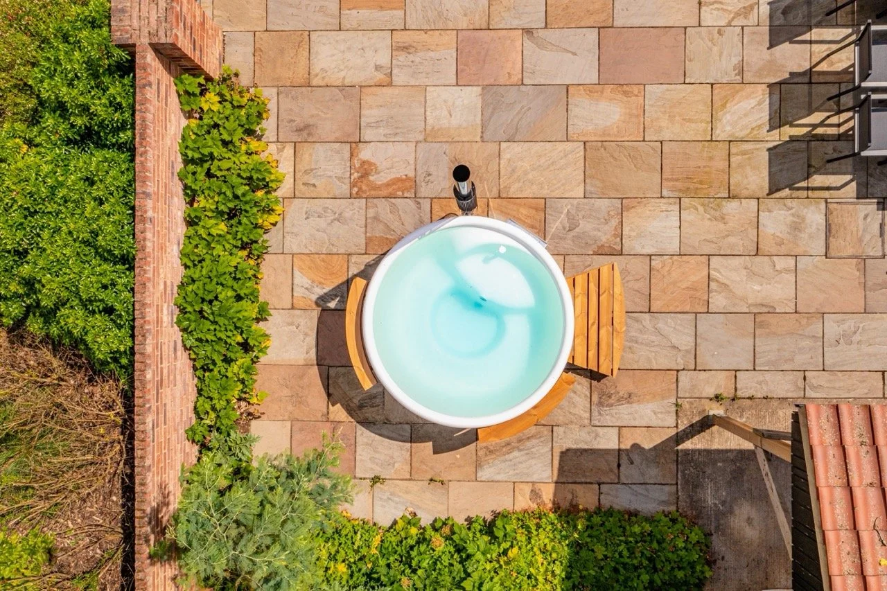 A top-down view of a hot tub with light blue water, surrounded by wooden seats, on a stone patio next to green plants and a brick wall.