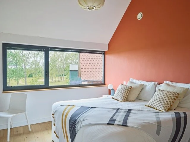 A bedroom with a large window showing trees and a building outside. The room has a bed with white linens and multiple pillows, a white chair, and an orange accent wall.
