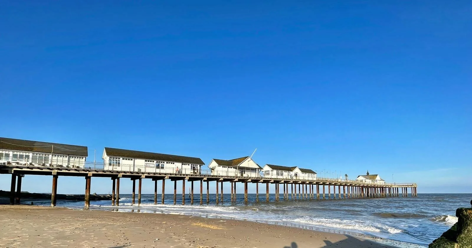 View of a beach with a wooden pier extending into the ocean, topped with small white buildings, under a clear blue sky.
