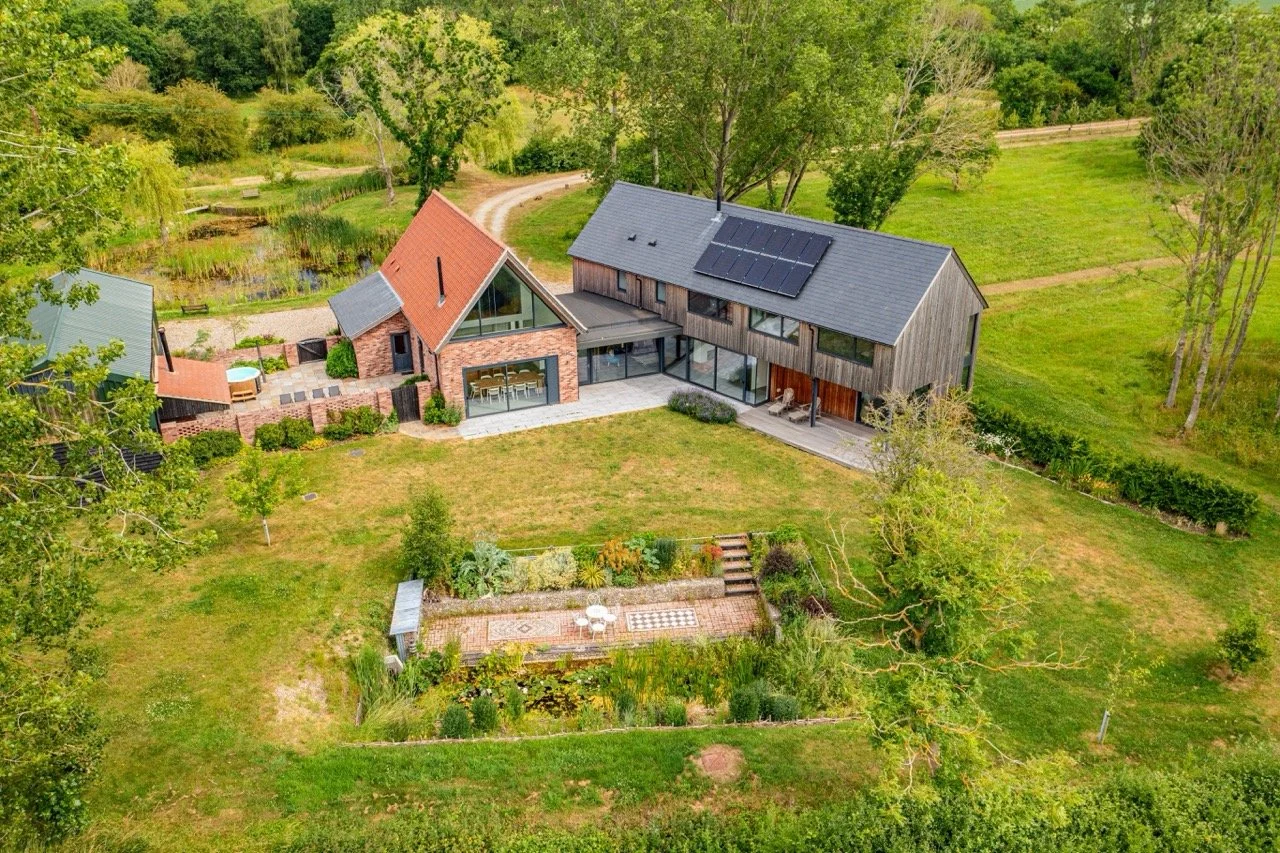 Aerial view of a modern house surrounded by green trees and a large grassy yard, with a garden and patio area in the backyard, and a pond nearby.