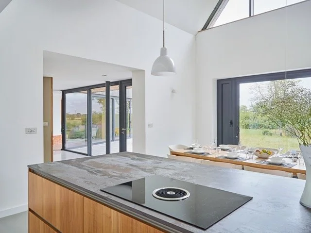 Modern kitchen with white walls, large windows, and a gray countertop on a wooden island. A dining table with dinnerware is visible near the window, and there is a white pendant light hanging from the ceiling.