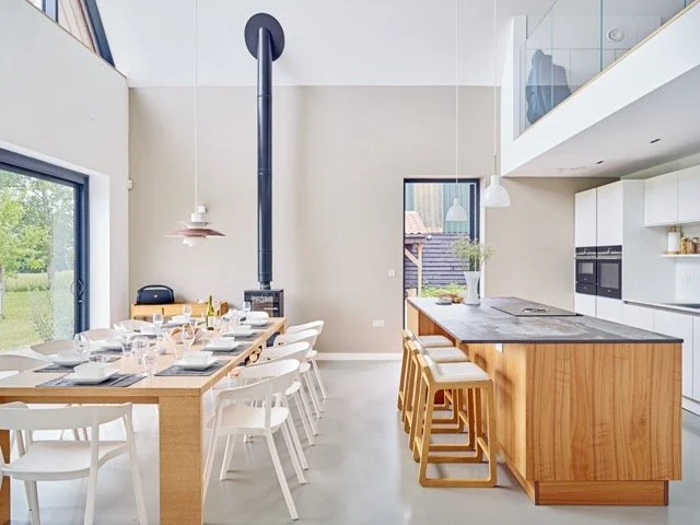 Modern dining area with a wooden table, white chairs, a kitchen island with bar stools, and large glass doors opening to an outdoor space.