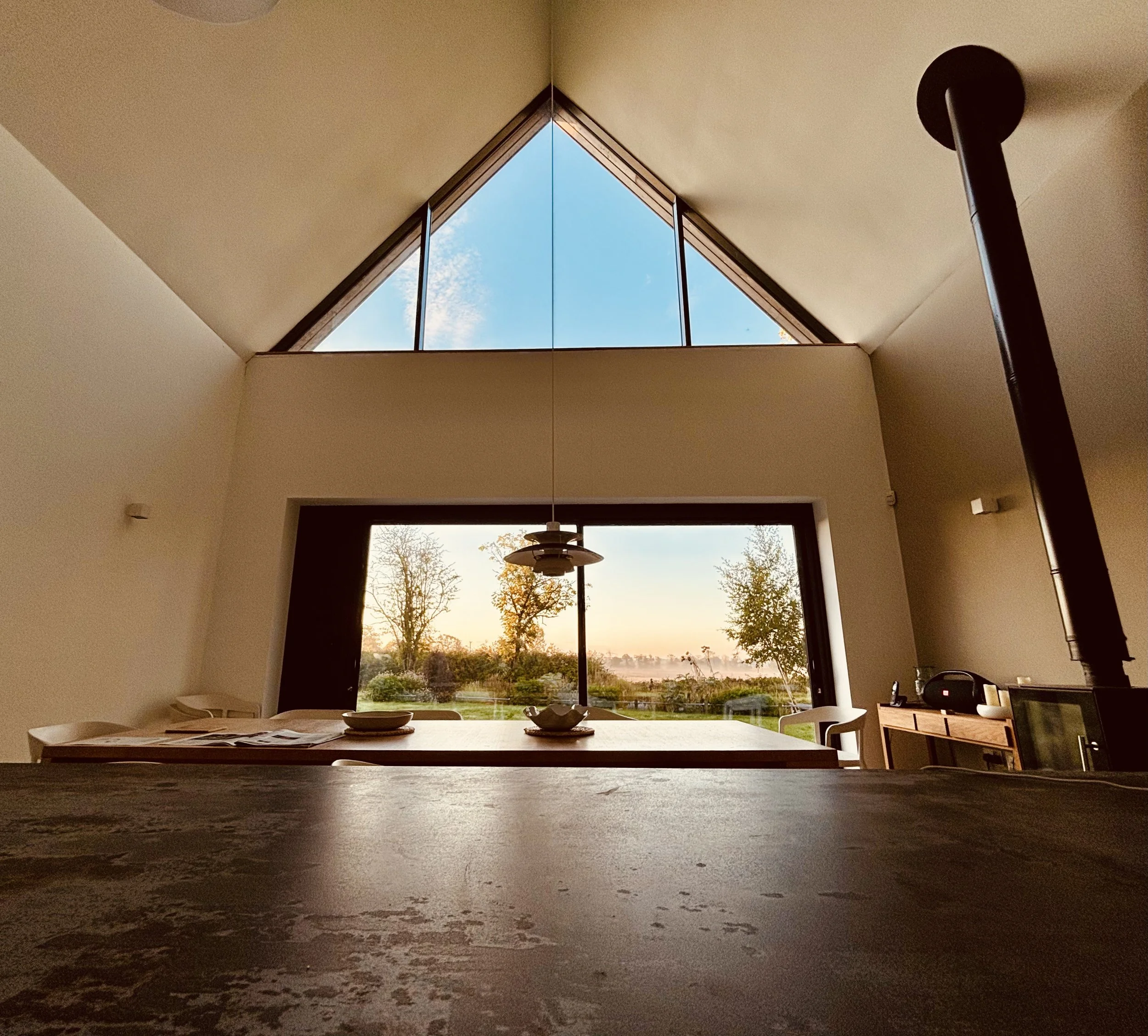 Interior of a modern room with a large window showing trees and a sunset, featuring a table with bowls and papers, a hanging pendant light, and a wood stove pipe.