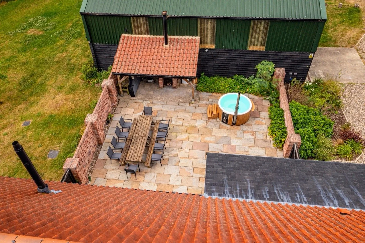 Aerial view of a backyard patio with outdoor furniture, a hot tub, Pizza oven and BBQ, a brick wall, garden beds, and adjacent buildings with roofs.