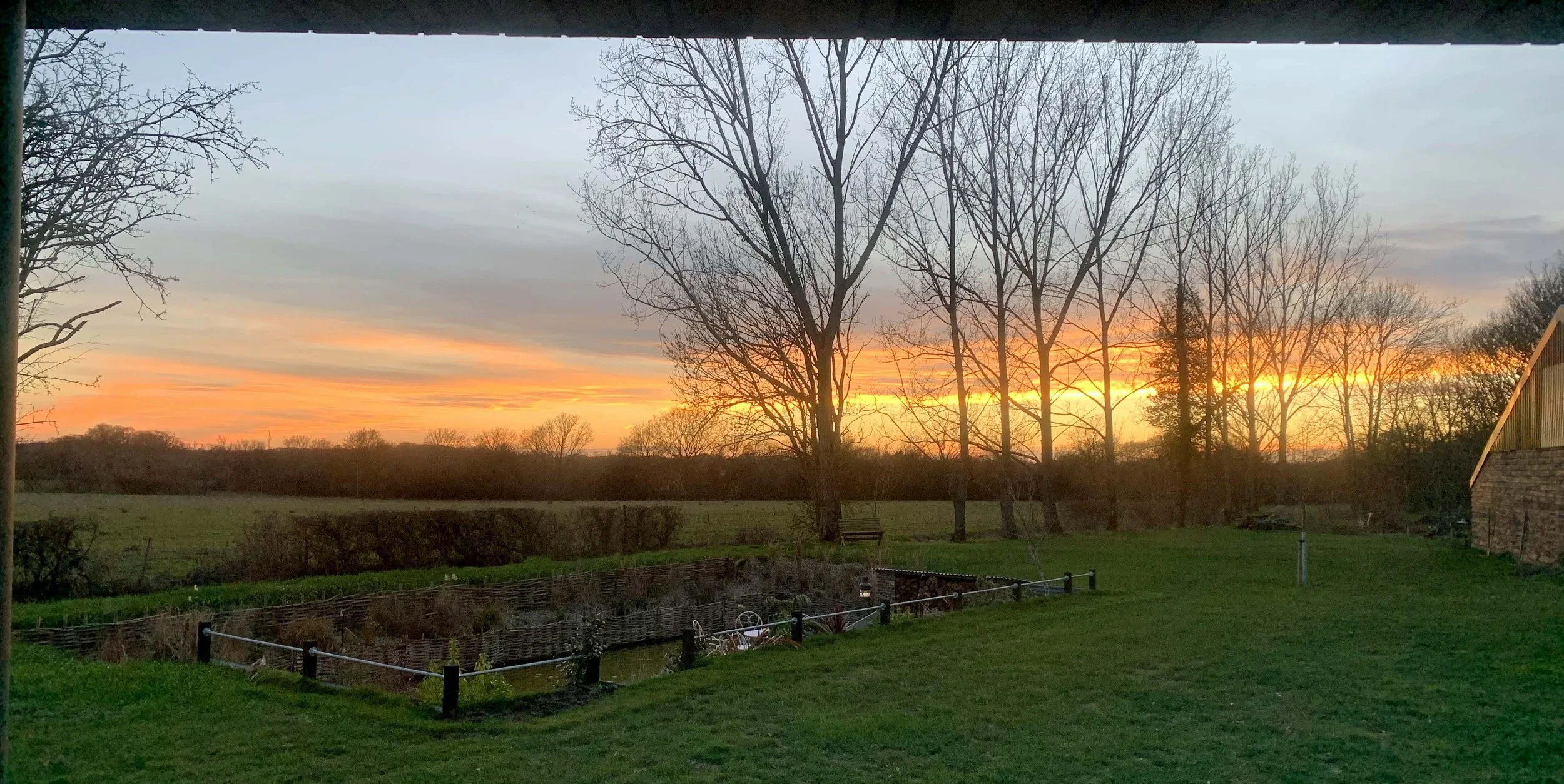A scenic sunrise over a rural landscape with leafless trees, green grass, and a stone building on the right. The sky is filled with orange, pink, and gray hues.