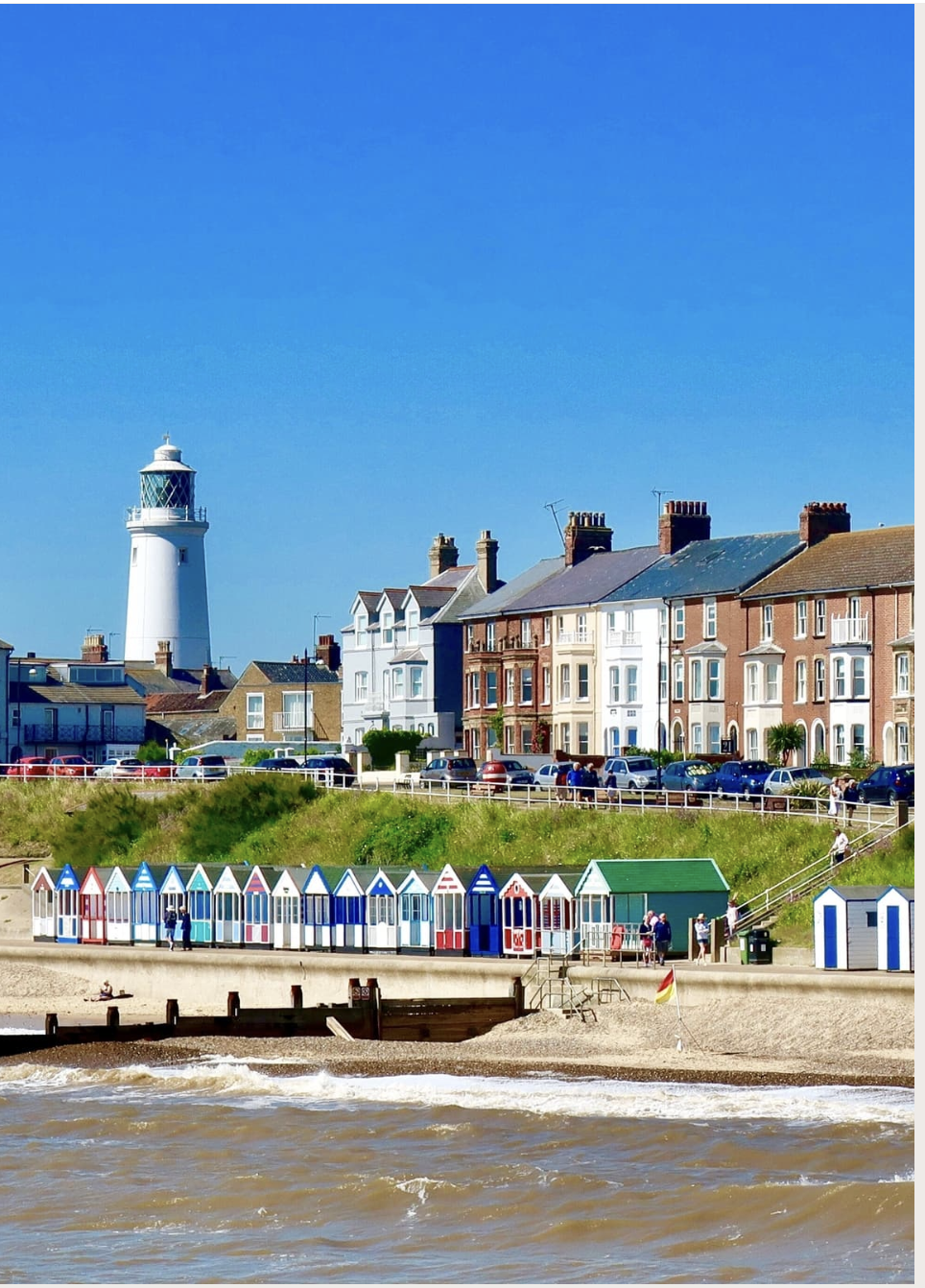 Colorful beach huts on a seaside promenade with a lighthouse and residential buildings in the background under a clear blue sky.