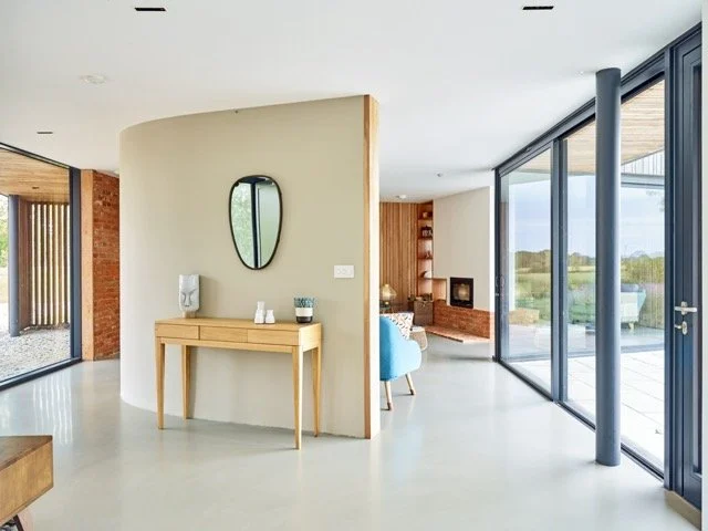 Modern living room with large floor-to-ceiling glass windows, a beige curved wall with a mirror, a wooden console table, and a view of an outdoor patio and green landscape.