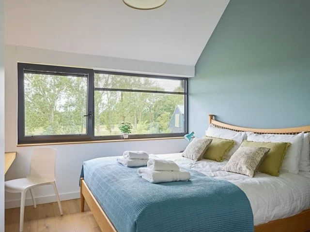 Bedroom with a large window, bed with white and green pillows, blue blanket, and towels, small potted plant on windowsill, light wood desk and white chair, green accent wall.
