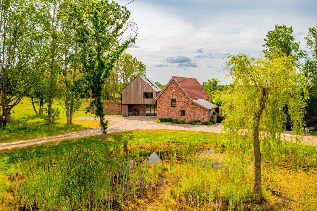 A rural house with a brick and wood exterior, surrounded by trees and a pond in the foreground, under a cloudy sky.