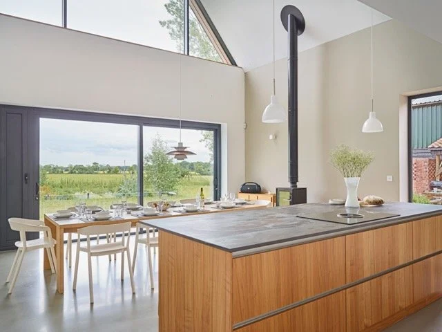 Modern dining area and kitchen with large window, wooden dining table set with plates and glasses, and a kitchen island with a vase and decorative items, in a bright open space.