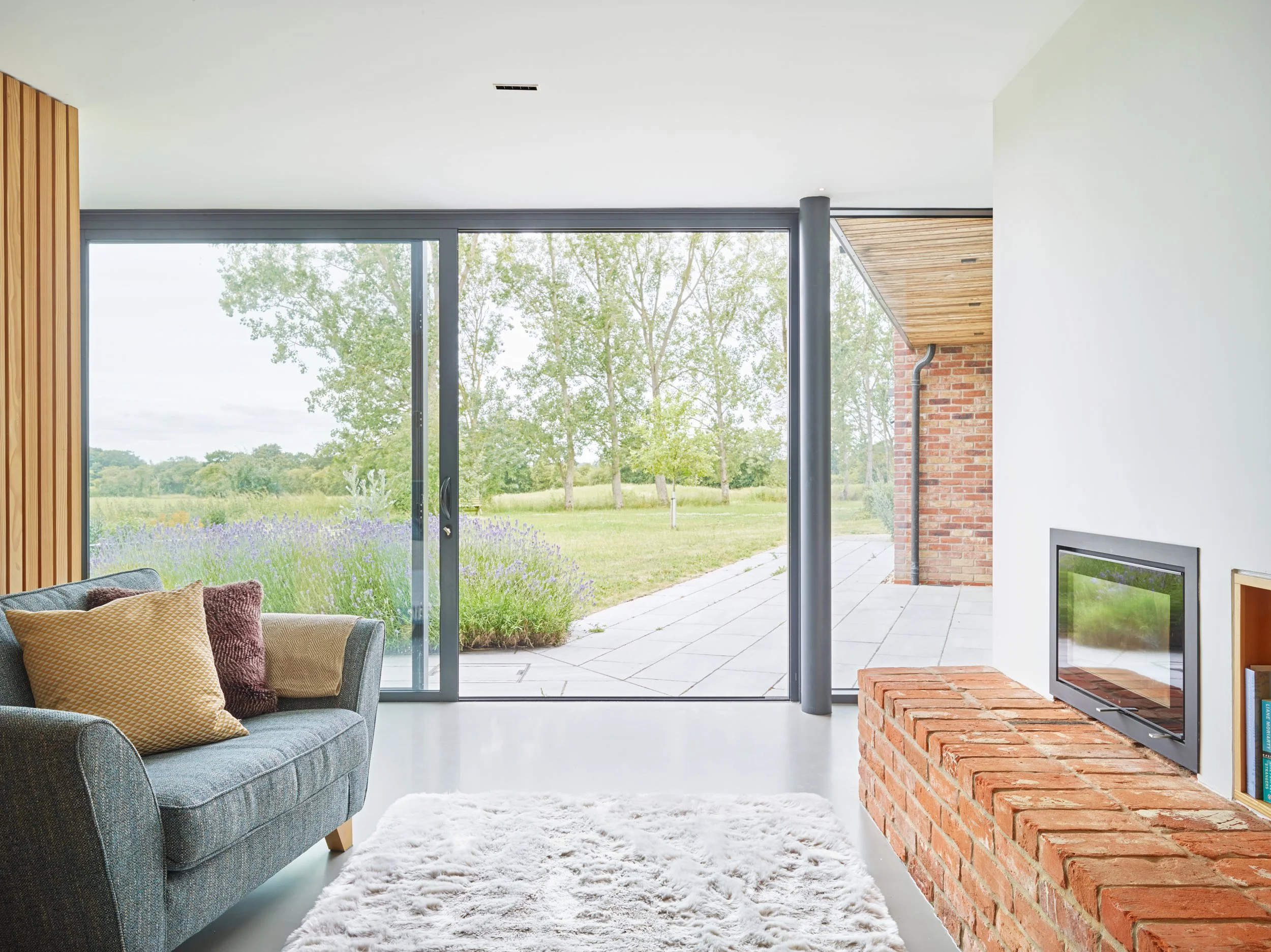 Modern living room with large sliding glass doors leading to a garden, a sofa with cushions, a white rug, a brick fireplace with built-in bookshelf, and a view of green trees and purple flowers outside.