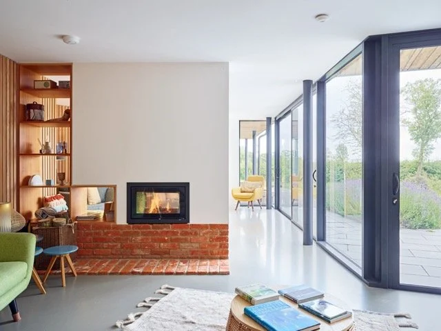Modern living room with a brick fireplace, wooden shelving on the left, and large glass sliding doors on the right showing an outdoor patio and greenery.