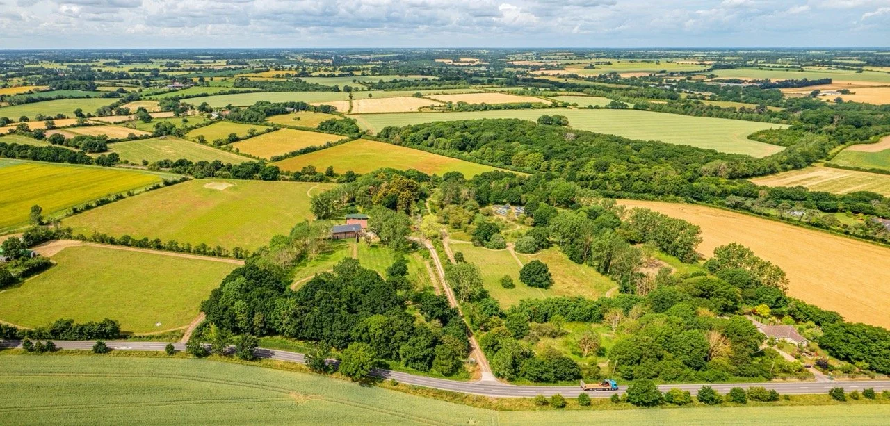 Aerial view of rural farmland with green fields, trees, and a few houses, under a partly cloudy sky.