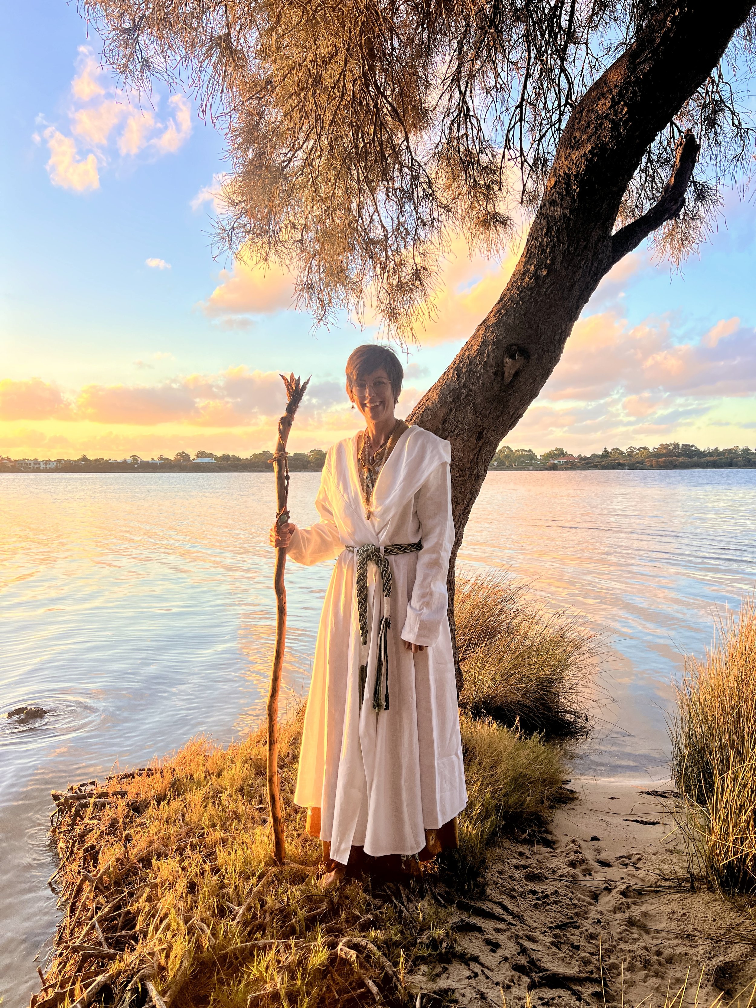 A druid woman, Charlie Bigwood dressed in white standing by a riverbank at sunset, holding a wooden staff, with a large tree behind her and colorful clouds in the sky.