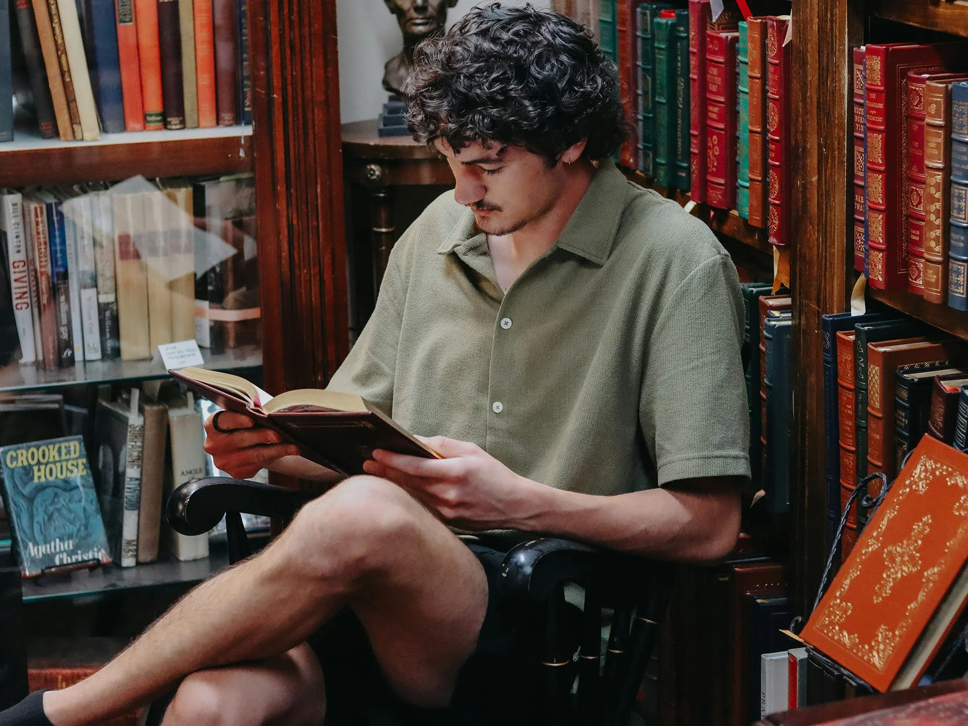 A young man with curly dark hair, light skin, and a small earring in his left ear is sitting in a bookstore or library, reading a book. He is wearing a greenish button-up shirt and is seated with one leg crossed over the other. Behind him are shelves filled with colorful books and a bust sculpture is visible on a table behind him.