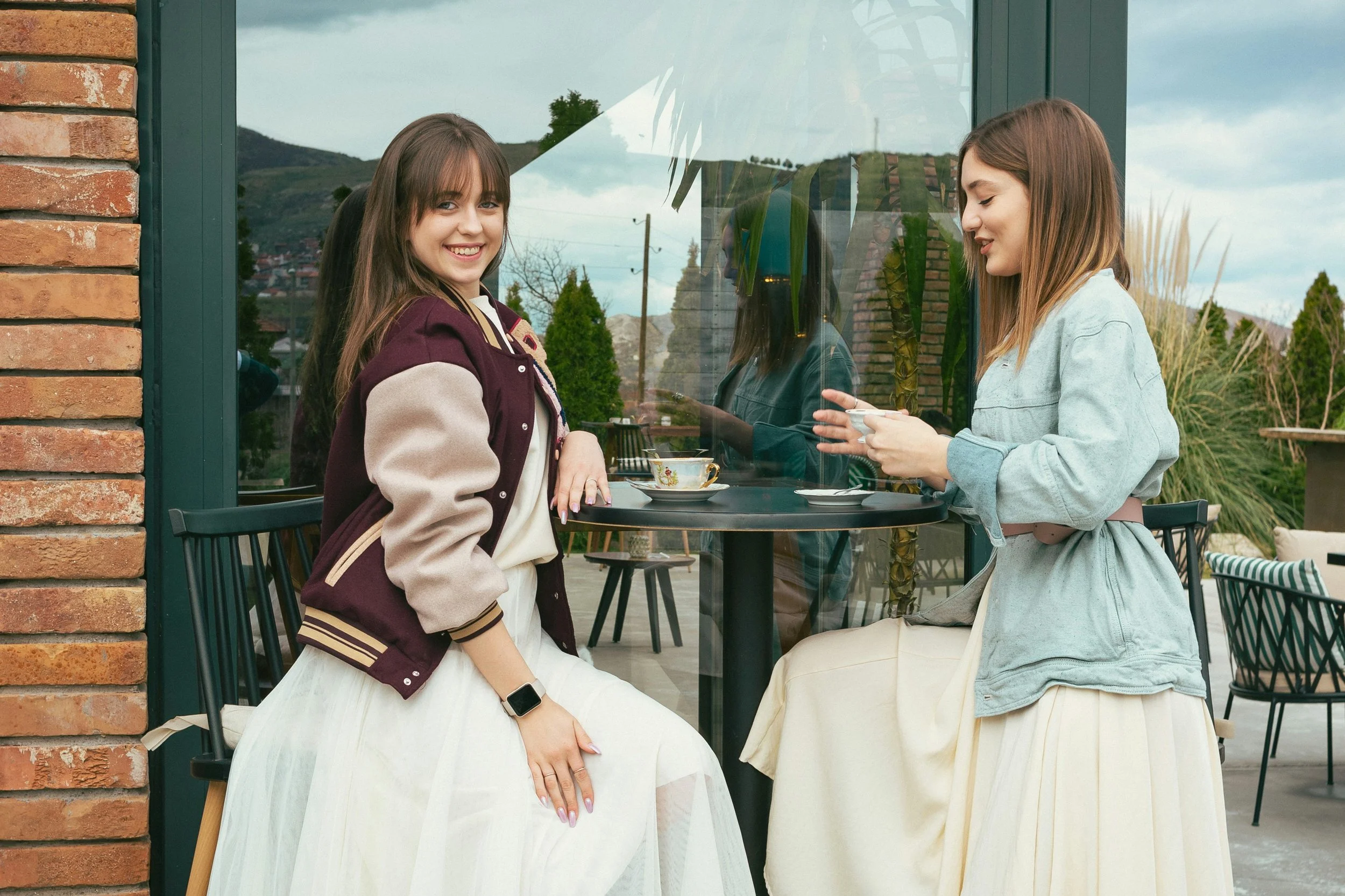 Two young women sitting outside near a building with a large window, engaged in conversation, with mountains and cloudy sky in the background.