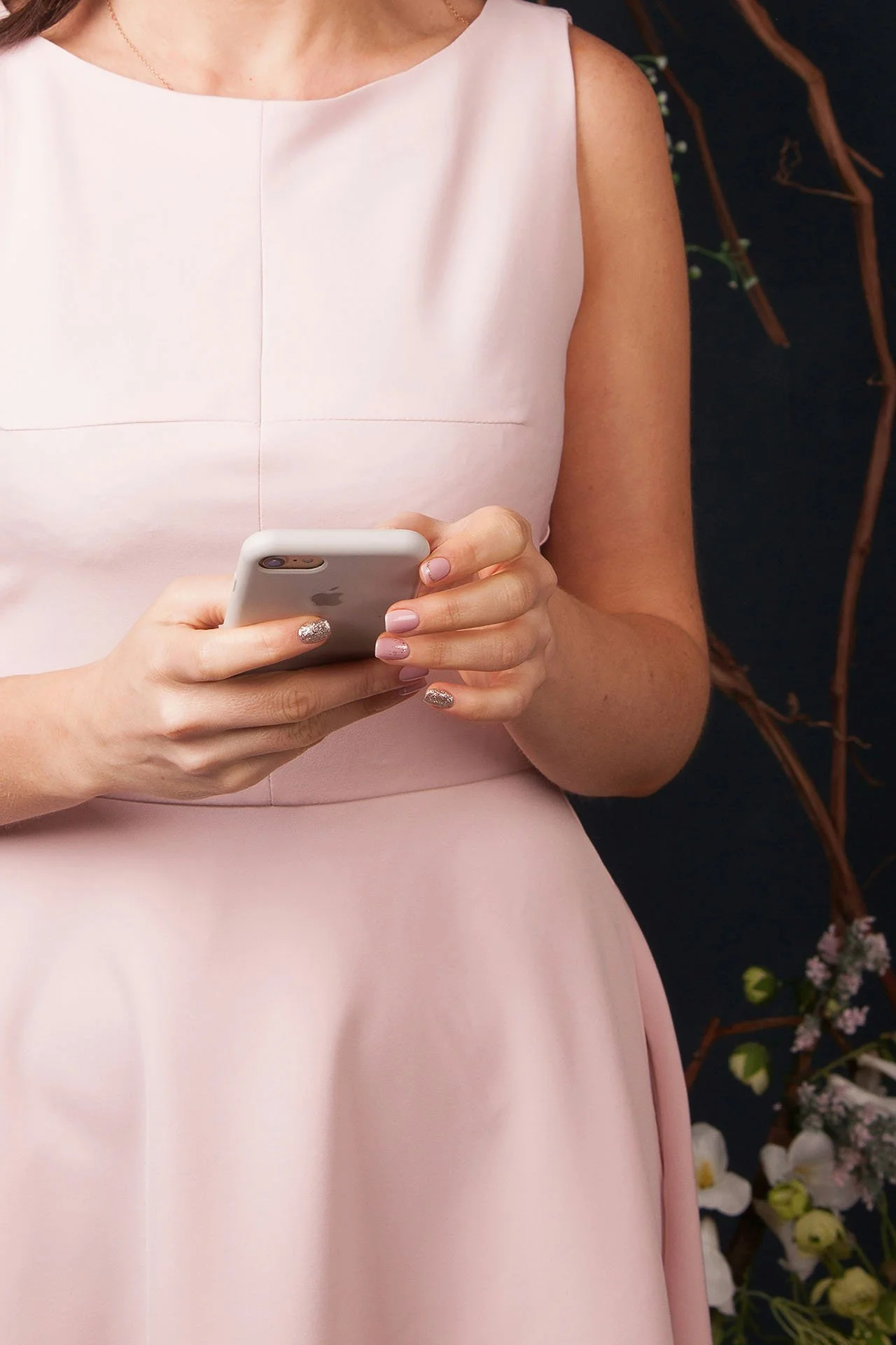 Close-up of a woman in a pink dress holding a smartphone, with manicured nails and jewelry, standing near floral decorations.