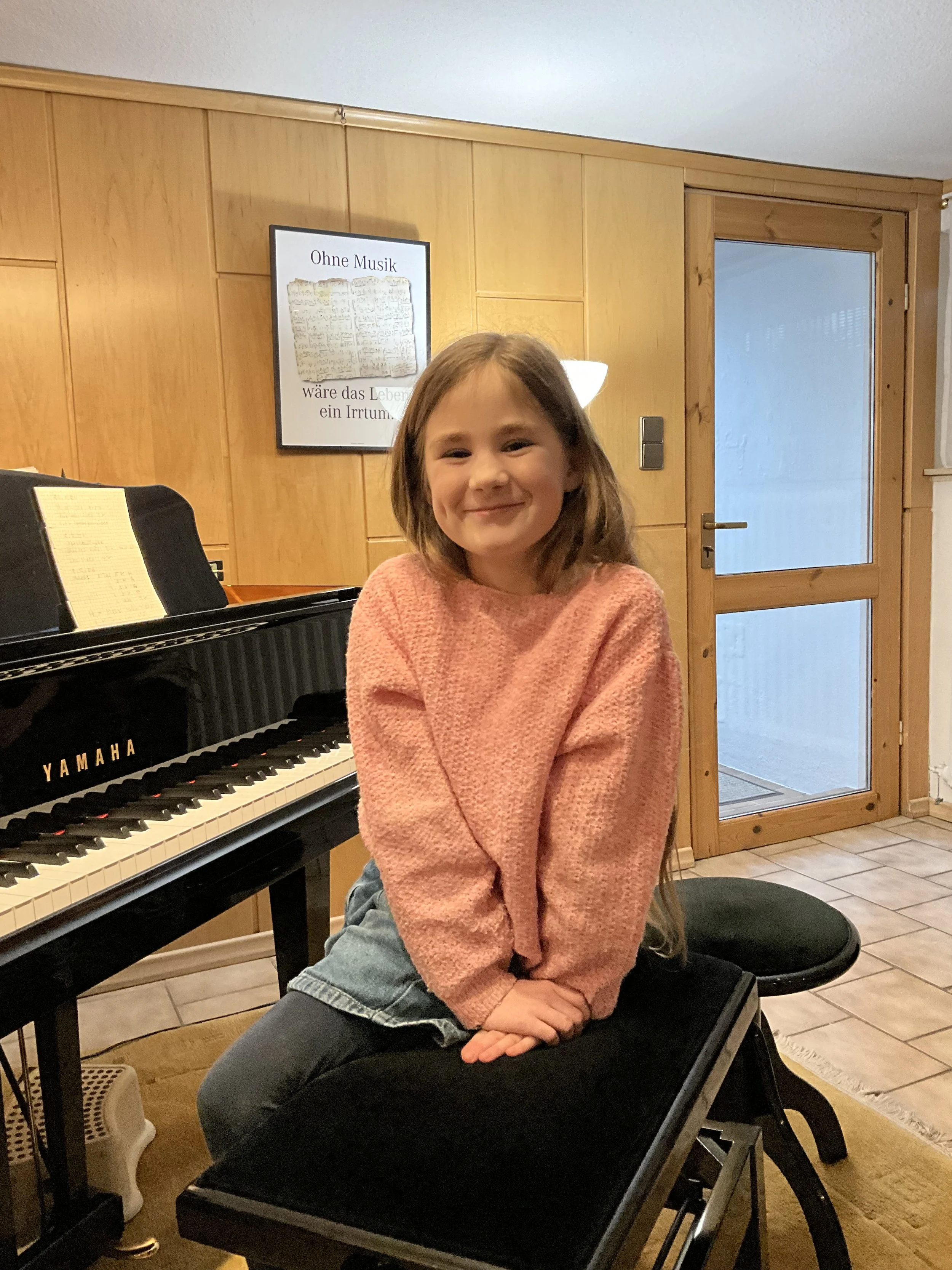 Ein lachendes Mädchen sitzt auf einer Klaviertastatur in einem Raum mit Holzwänden und einer Glastür im Hintergrund.