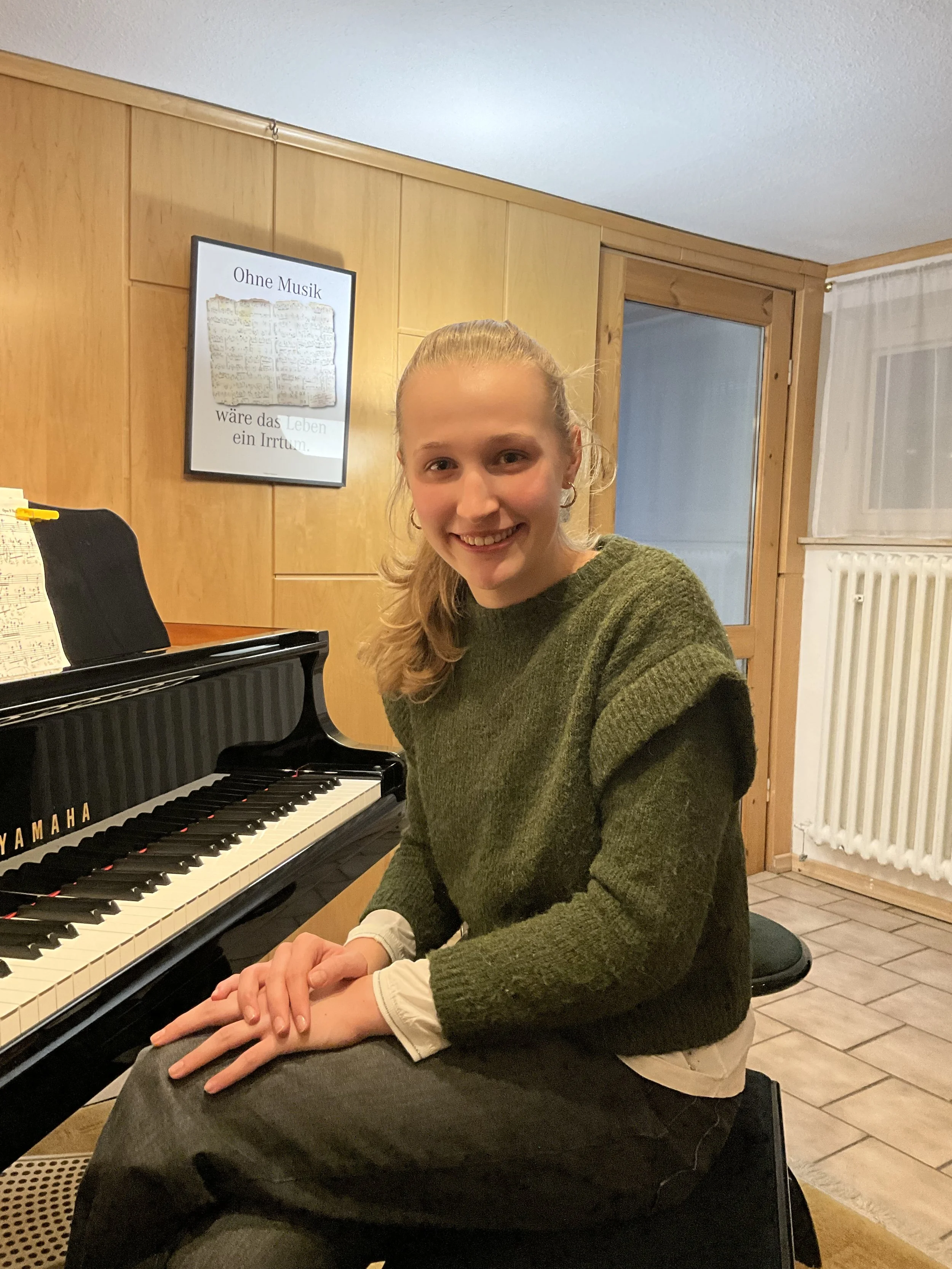 Junge Frau spielt Klavier und lächelt im Raum mit Holzverkleidung und einem Poster an der Wand, das den Titel 'Ohne Musik wäre das Leben ein Irrtum' trägt.