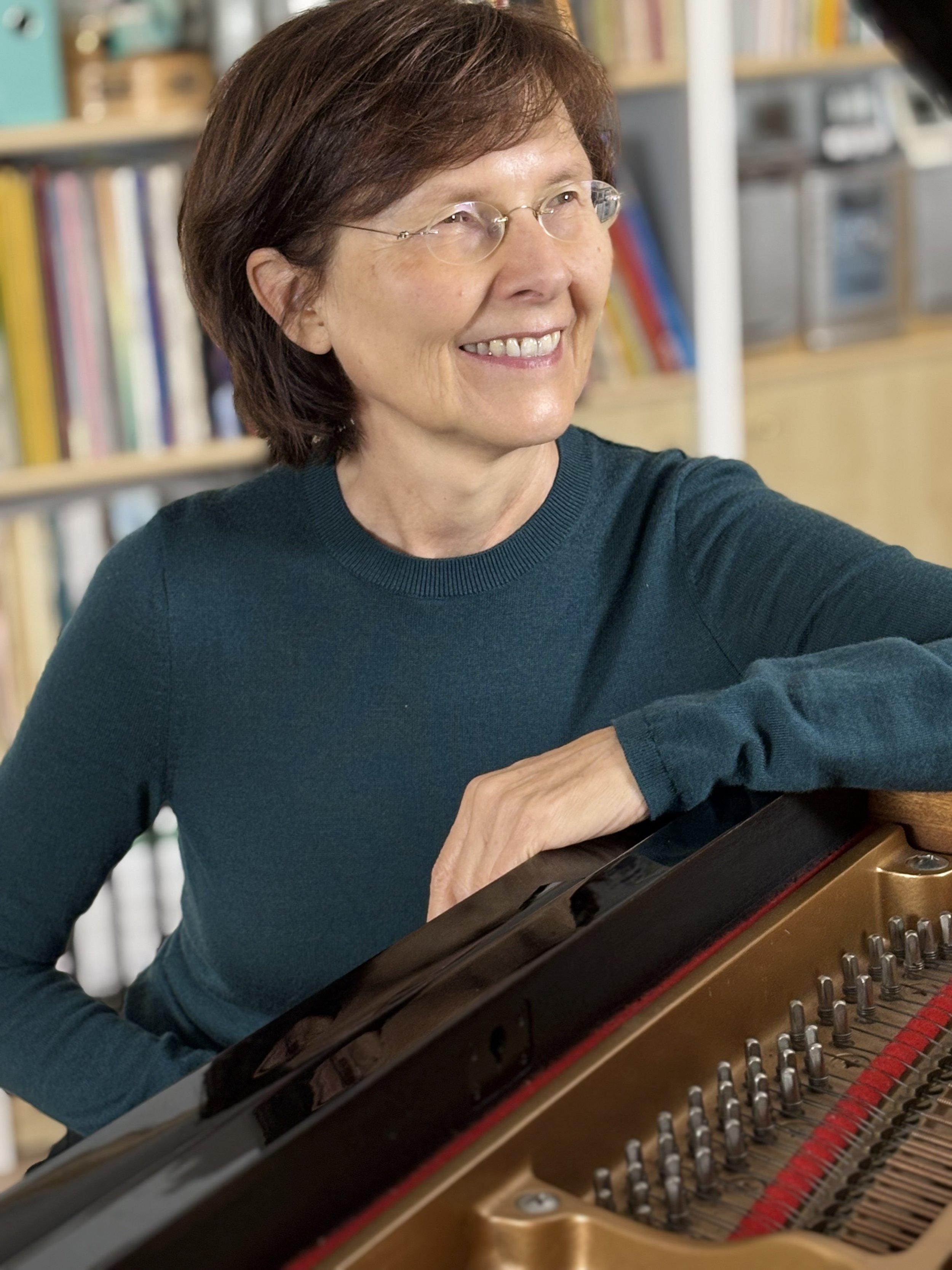 Beate May mit Brille spielt Klavier in einem Raum mit Büchern im Hintergrund.
