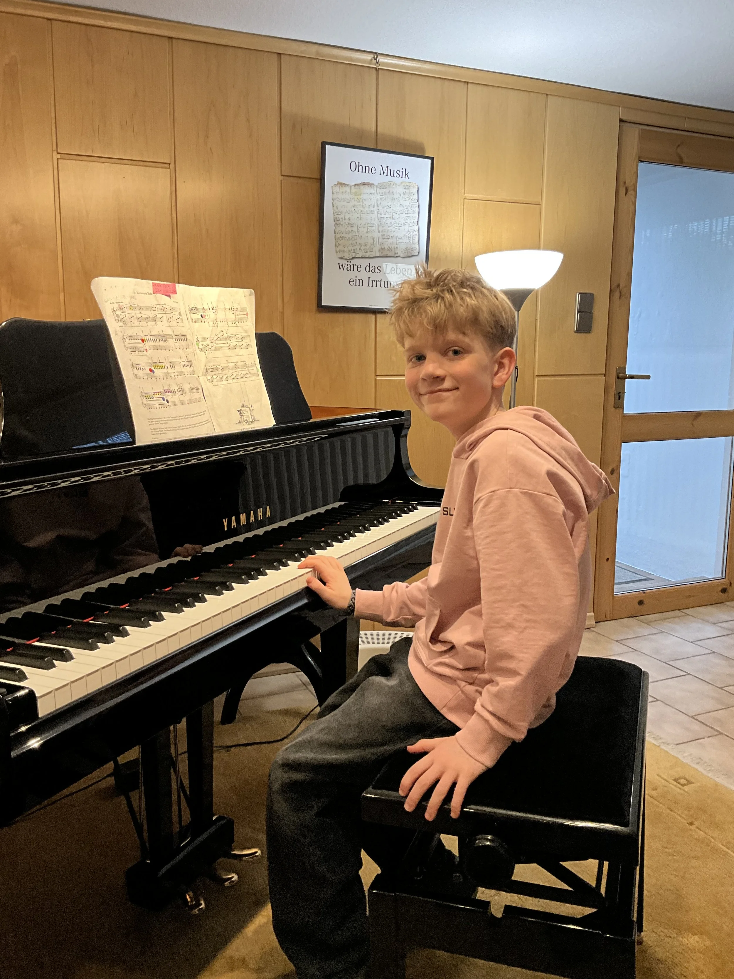 Ein Junge sitzt an einem Flügel mit aufgeschlagenem Notenblatt, im Hintergrund eine Stehlampe und eine Holzwand. Auf dem Bild steht ein Rahmen mit einem Bild und deutschem Text.