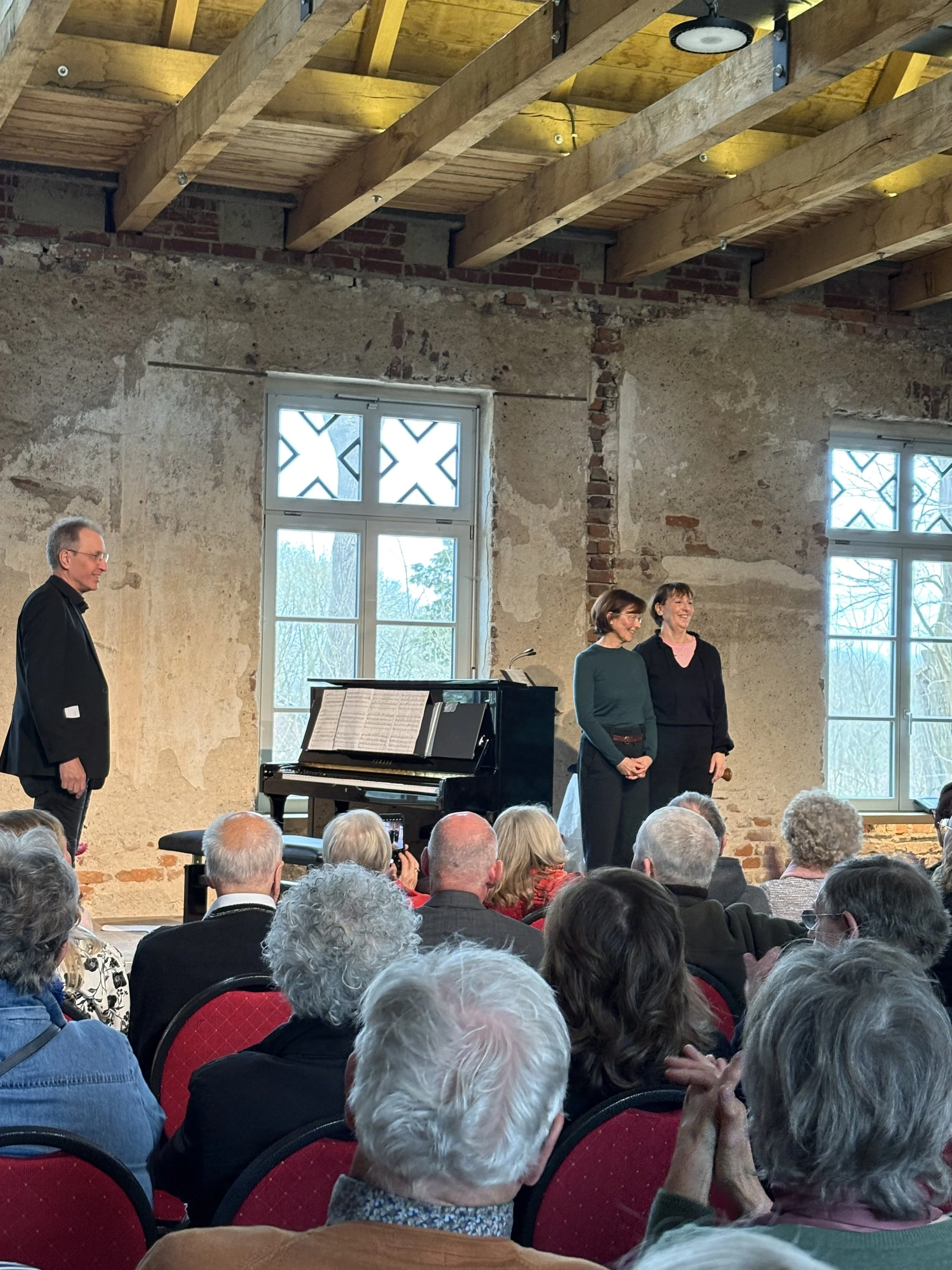 Ein Konzert- oder Theaterveranstaltung in einem Raum mit alter Wandgestaltung und großen Fenstern. Es sind mehrere Menschen im Publikum, zwei Frauen stehen auf der Bühne neben einem Klavier, ein Mann steht auf der anderen Seite der Bühne.