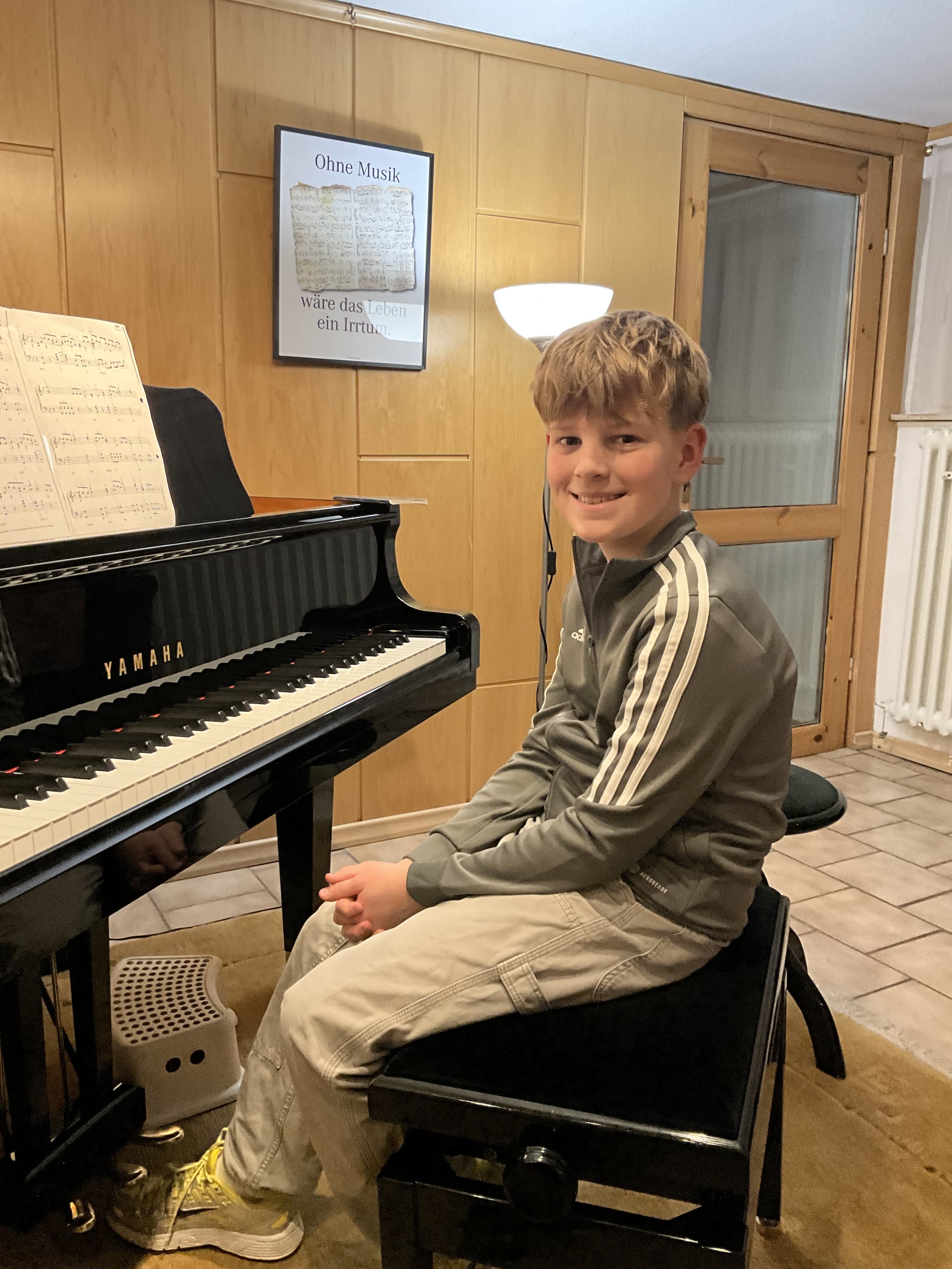 Junge sitzt an einem Klavier in einem Raum mit Holzvertäfelung, lächelt zur Kamera, Hintergrund mit Wandlampe, Fenster und einem Bild mit Text.