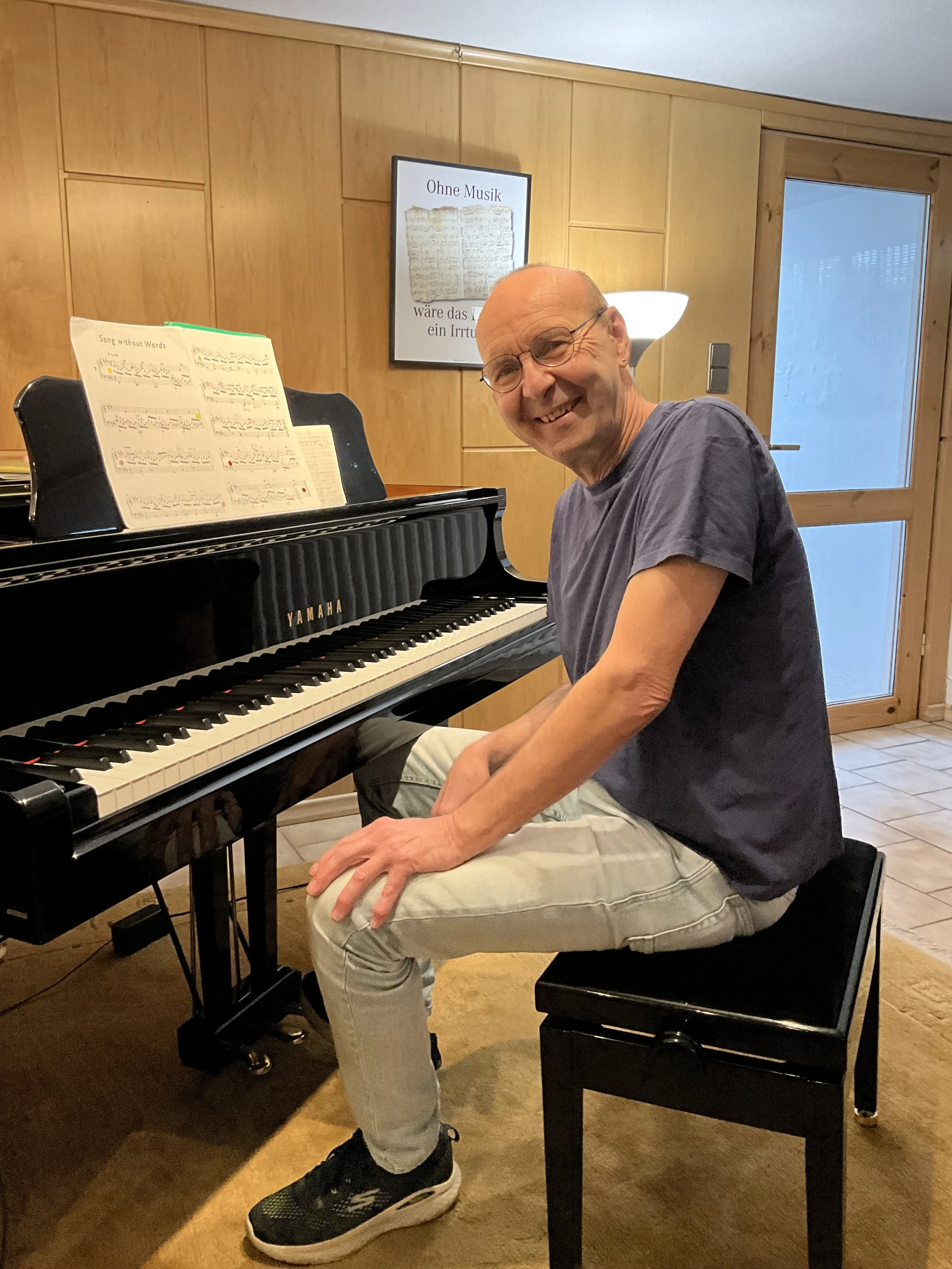 Ein lachender Mann sitzt am Klavier in einem Raum mit Holzvertäfelung an der Wand. Ein Notenblatt ist auf dem Klavier, und im Hintergrund ist ein Bild mit dem Titel 'Ohne Musik wäre das Leben ein Irrtum' zu sehen.