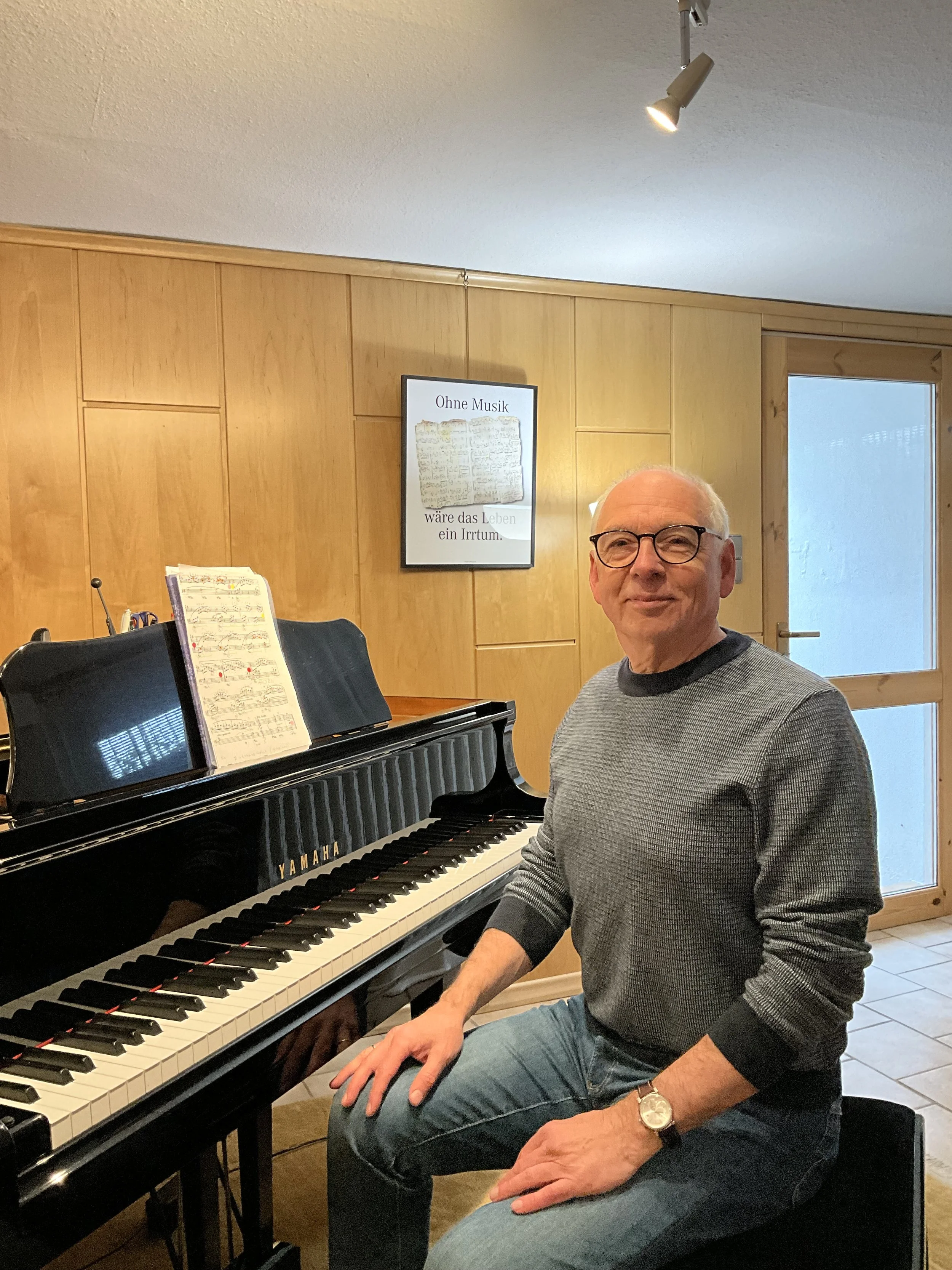 Ein älterer Mann mit Brille sitzt am Klavier in einem Raum mit Holzverkleidung. Es gibt ein Bild an der Wand mit einem Zitat auf Deutsch.
