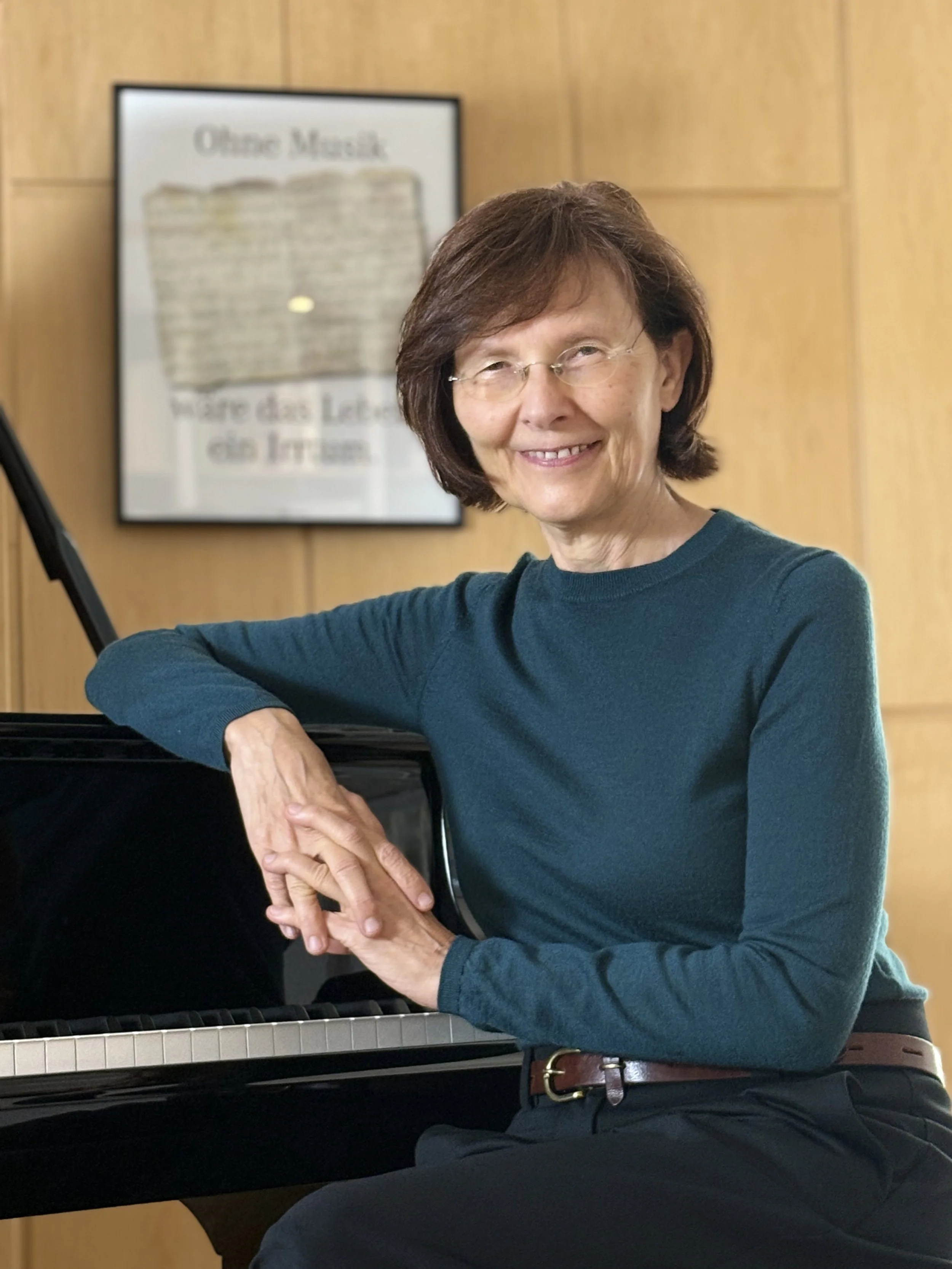 Beate May sitzt an einem Klavier mit einer Hand auf der Klaviatur, vor einem Holzpaneel Wand, im Hintergrund ein gerahmtes Musikposter.